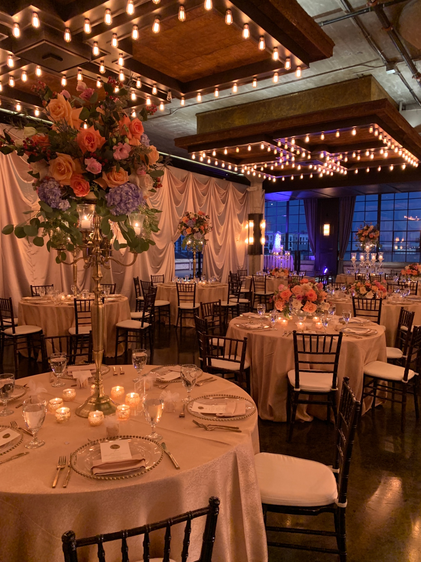Warm candlelit reception with tall coral and lavender floral candelabras under Edison-bulb canopy