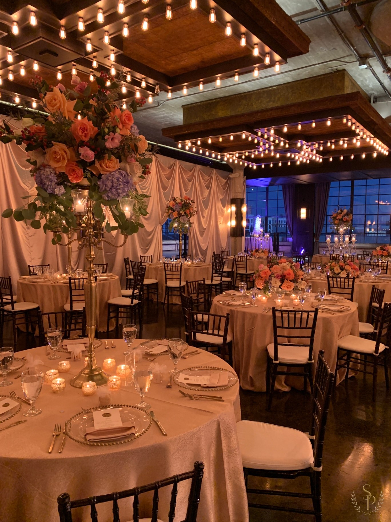 Warm candlelit reception with tall coral and lavender floral candelabras under Edison-bulb canopy